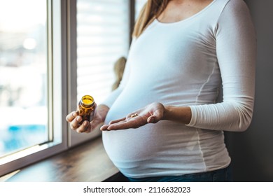 Pregnant Woman Reading Label On Bottle With Medicine, With Vitamins. Female Sitting At Home In Bed With Glass Of Water Medicine. Pregnancy, Health, Pharmaceuticals, Care And People.