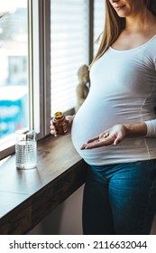 Pregnant Woman Reading Label On Bottle With Medicine, With Vitamins. Female Sitting At Home In Bed With Glass Of Water Medicine. Pregnancy, Health, Pharmaceuticals, Care And People.