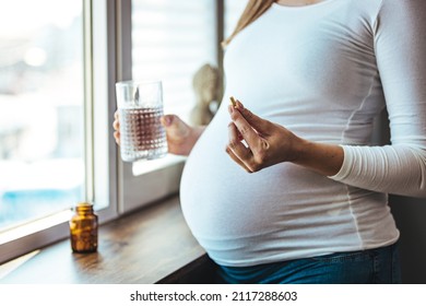 Pregnant woman reading label on bottle with medicine, with vitamins. Female sitting at home in bed with glass of water medicine. Pregnancy, health, pharmaceuticals, care and people. - Shutterstock ID 2117288603