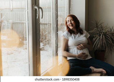 Pregnant Woman With Red Hair Laughs While Sitting At Home By The Window. Happy Pregnancy, Real Life Of A Pregnant Lady. High Quality Photo