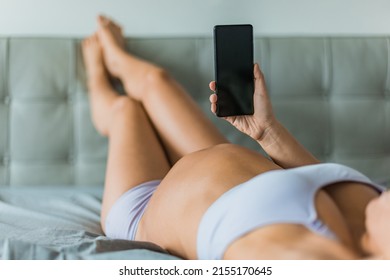 Pregnant Woman Relaxing In Bed Using Phone App For Online Pregnancy Prenatal Class. Focus On Mobile And Baby Bump Belly, Girl Lying On Back In Comfortable Underwear