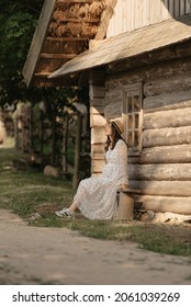 A Pregnant Woman Is Relaxing Near The Old Log House In An Eastern European Village. A Happy Pregnancy. A Lady In A Long Dress And A Hat Sitting On A Bench In The Countryside In The Evening.