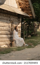 A Pregnant Woman Is Relaxing Near The Old Log House In An Eastern European Village. A Happy Pregnancy. A Lady In A Long Dress And A Hat Sitting On A Bench In The Countryside In The Evening.