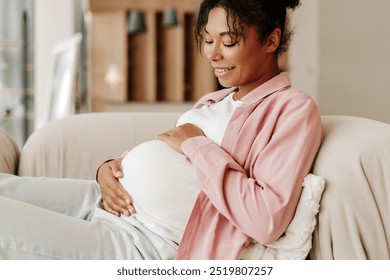 Pregnant woman is relaxing on a sofa at home, gently holding her belly and smiling - Powered by Shutterstock