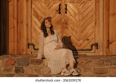 A Pregnant Woman Is Relaxing On The Stone Steps Of The Old Church In An Eastern European Village. A Happy Pregnancy. A Lady In A Long Dress And A Hat Sitting In The Countryside In The Evening.