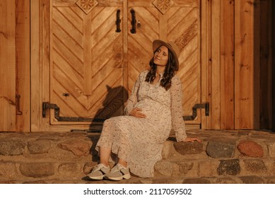 A Pregnant Woman Is Relaxing On The Stone Steps Of The Old Church In An Eastern European Village. A Happy Pregnancy. A Lady In A Long Dress And A Hat Sitting In The Countryside In The Evening.