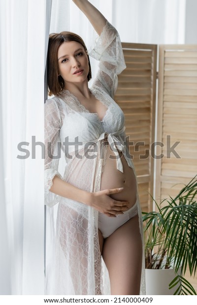 Pregnant woman in robe posing near curtains and\
plant at home