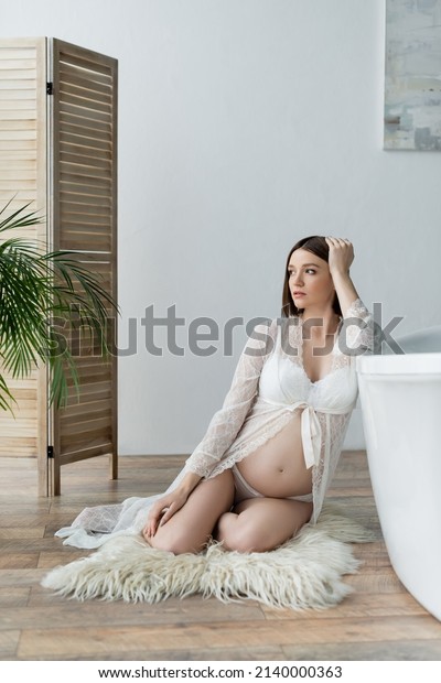 Pregnant woman in robe sitting on fluffy rug near\
bathtub at home