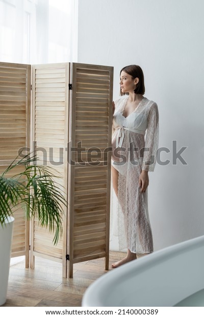 Pregnant woman in robe standing near folding screen\
and bathtub at home