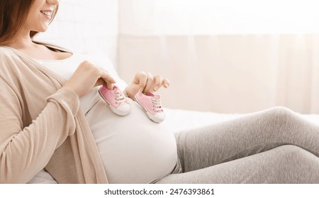 A pregnant woman is seated on a couch, holding a pink baby shoe in her hand. She appears contemplative as she gazes at the tiny shoe, possibly imagining her future with her soon-to-arrive baby. - Powered by Shutterstock