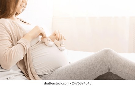 A pregnant woman is seen resting comfortably on bed, holding a pair of baby shoes near her belly. She appears content and serene, enjoying a quiet moment, cropped - Powered by Shutterstock