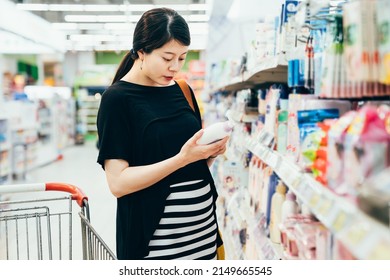 Pregnant Woman Shopping At Cosmetic Department In Wholesale Store. Elegant Asian Japanese Mom With Big Belly Holding Shampoo Product And Reading Ingredient To Protect Baby In Abdomen In Supermarket