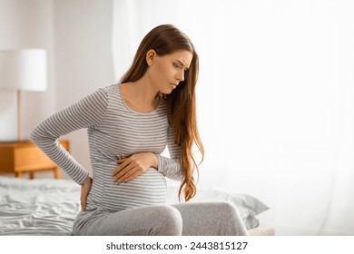 A pregnant woman shows signs of discomfort or cramps, sitting on the bed with a worried expression - Powered by Shutterstock