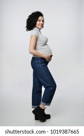 Pregnant Woman Side View Standing In Full Length Smiling Holding Touching Her Tummy Looking At It, Over Grey Background