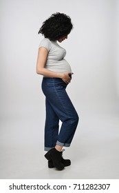 Pregnant Woman Side View Standing In Full Length Smiling Holding Touching Her Tummy Looking At It, Over Grey Background