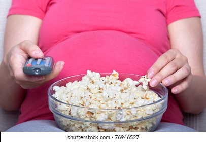 Pregnant woman sit and eat popcorn on sofa, watching television