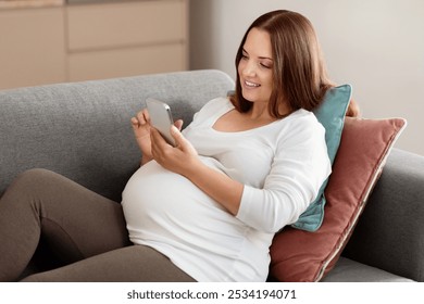 A pregnant woman sits comfortably on a couch, enjoying her time as she scrolls through her phone, surrounded by soft pillows in a cozy living space. - Powered by Shutterstock