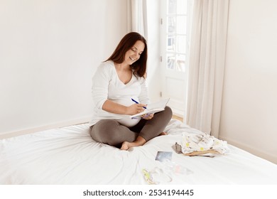 A pregnant woman sits cross-legged on a bed, happily writing in a notebook. She is surrounded by baby clothes and enjoys a moment of creativity in a well-lit, airy room. - Powered by Shutterstock