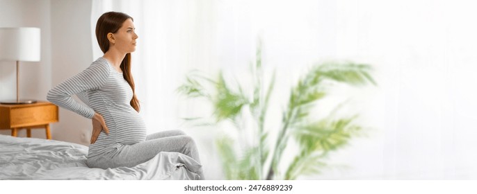 A pregnant woman sits on the edge of her bed in her bedroom, holding her back in pain. She appears to be experiencing discomfort, possibly related to pregnancy, copy space - Powered by Shutterstock