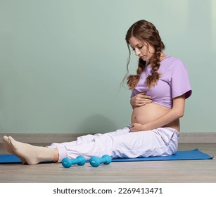 A pregnant woman sits on fitness mat. Female looks at her pregnant belly. - Shutterstock ID 2269413471