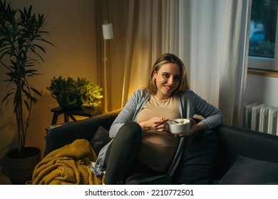 Pregnant Woman Sits On Sofa In Living Room, Eats Fruit Salad And Looking At Camera