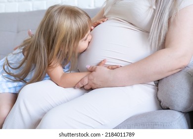 Pregnant Woman Sitting On Bed With Little Baby Daughter Stroking And Kissing Tummy