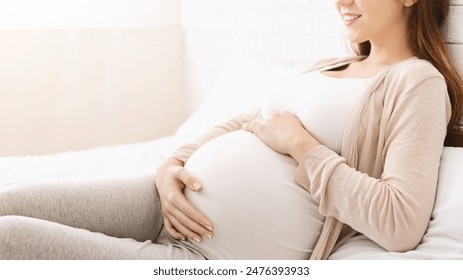 A pregnant woman is sitting on a bed with a smile on her face. The room is simple and uncluttered, emphasizing the woman joyful expression, cropped, copy space - Powered by Shutterstock