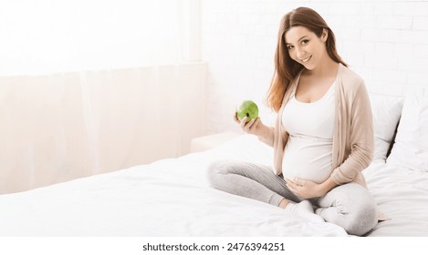 A pregnant woman is sitting on a bed while holding a green apple. She appears relaxed and content as she enjoys a healthy snack. The woman baby bump is visible, highlighting her pregnancy journey. - Powered by Shutterstock