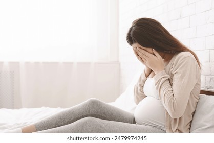 A pregnant woman sitting on a bed with her hands covering her face. She appears to be in a moment of contemplation or distress. - Powered by Shutterstock