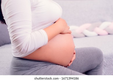 Pregnant woman sitting on the couch and holding her pregnant baby with two hands wearing withe top. Motherhood time. 