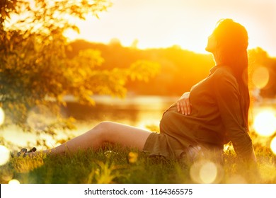 Pregnant Woman Sitting On Green Grass, Looking On River On Sunset, Touching Her Belly And Dreaming. Enjoying Healthy Pregnancy. Pregnant Tummy. Beauty Expectant Girl Outdoor In Summer Park In Sun Glow