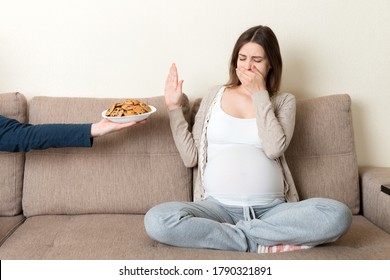 Pregnant woman sitting on the sofa refuses to eat cookies and makes no gesture. Healthy diet during pregnancy concept.