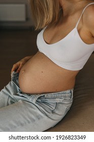 Pregnant Woman Sitting On A Sofa At Home, Wearing White Crop Top And Jeans, Closeup.