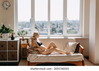 Pregnant Woman Sitting On A Sofa In Soft Light By The Window At Home, Wearing A Cozy Cardigan.