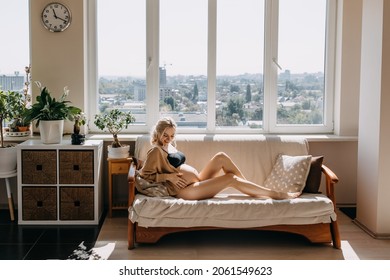 Pregnant Woman Sitting On A Sofa In Soft Light By The Window, Wearing A Cozy Cardigan.