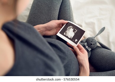 pregnant woman sitting on white bed and holds ultrasound baby
