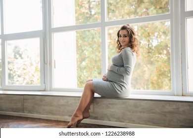 A Pregnant Woman Sitting On The Windowsill Near The Window At Home. She Dressed In Beautiful Maternity Dress