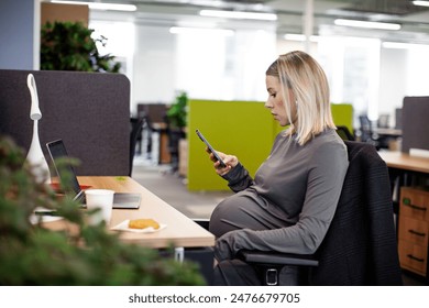 Pregnant woman sitting and working at a desk with a mobile phone in an office, with a computer and snacks on the table. - Powered by Shutterstock