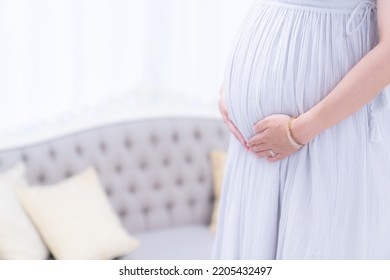 A Pregnant Woman Standing With Her Hands On Her Stomach In A Maternity Dress Indoors (shooting A Real Pregnant Woman)