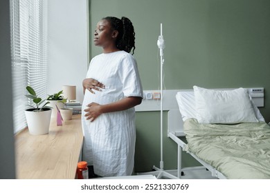 Pregnant woman standing near window in hospital room while looking outside. She appears contemplative, her hand placed gently on her belly, with hospital bed and potted plants nearby - Powered by Shutterstock