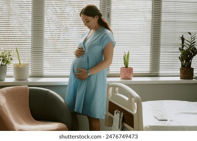 Pregnant woman standing next to window in hospital ward and caressing her belly - Powered by Shutterstock