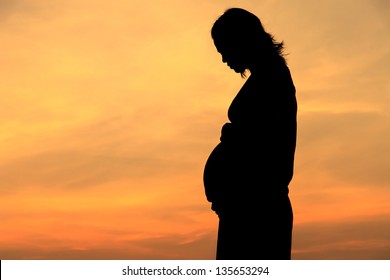 Pregnant woman standing on a beach by the sea stood backlit.