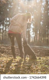 Pregnant Woman Standing In Sunlit Autumn Forest Reaching Down To Pet Her Black Dog Loyally Looking At Her.