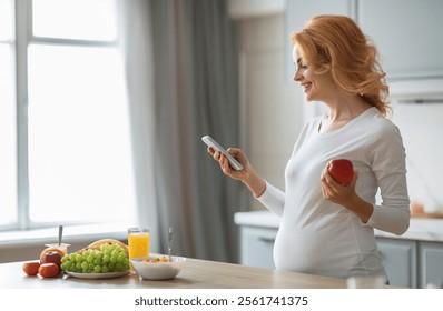 A pregnant woman stands in a modern kitchen, holding an apple in one hand and her phone in the other. A colorful spread of fresh fruits and juice is on the counter, emphasizing her healthy lifestyle. - Powered by Shutterstock