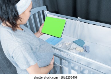 A Pregnant Woman Stands Near A Crib And Uses A Digital Tablet With A Green Screen For Chromakey Consults Online, Empty Space