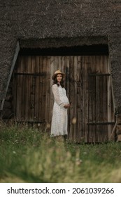 A Pregnant Woman Is Staying Near The Wooden Barn With A Cane Roof In An Eastern European Village. A Happy Pregnancy On The Farm. A Lady In A Long Dress And A Hat In The Countryside In The Evening.