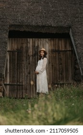 A Pregnant Woman Is Staying Near The Wooden Barn With A Cane Roof In An Eastern European Village. A Happy Pregnancy On The Farm. A Lady In A Long Dress And A Hat In The Countryside In The Evening.