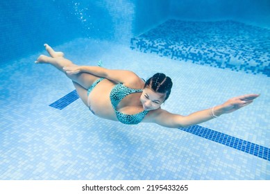 Pregnant Woman Swim, Dive In Swimming Pool. Aqua Fitness Healthcare Classes For Pregnancy. Underwater Picture Of Pregnant Woman Wearing Fashion Swimsuit In Blue Deep Pool. Aquatic Therapy Workout.