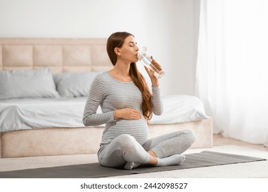 A pregnant woman takes a healthy sip of water while sitting cross-legged, highlighting the importance of hydration - Powered by Shutterstock