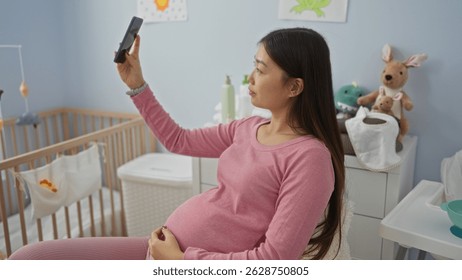 Pregnant woman taking selfie in cozy bedroom with baby cradle, showcasing her long dark hair and wearing a pink outfit, surrounded by soft toys and pastel decor